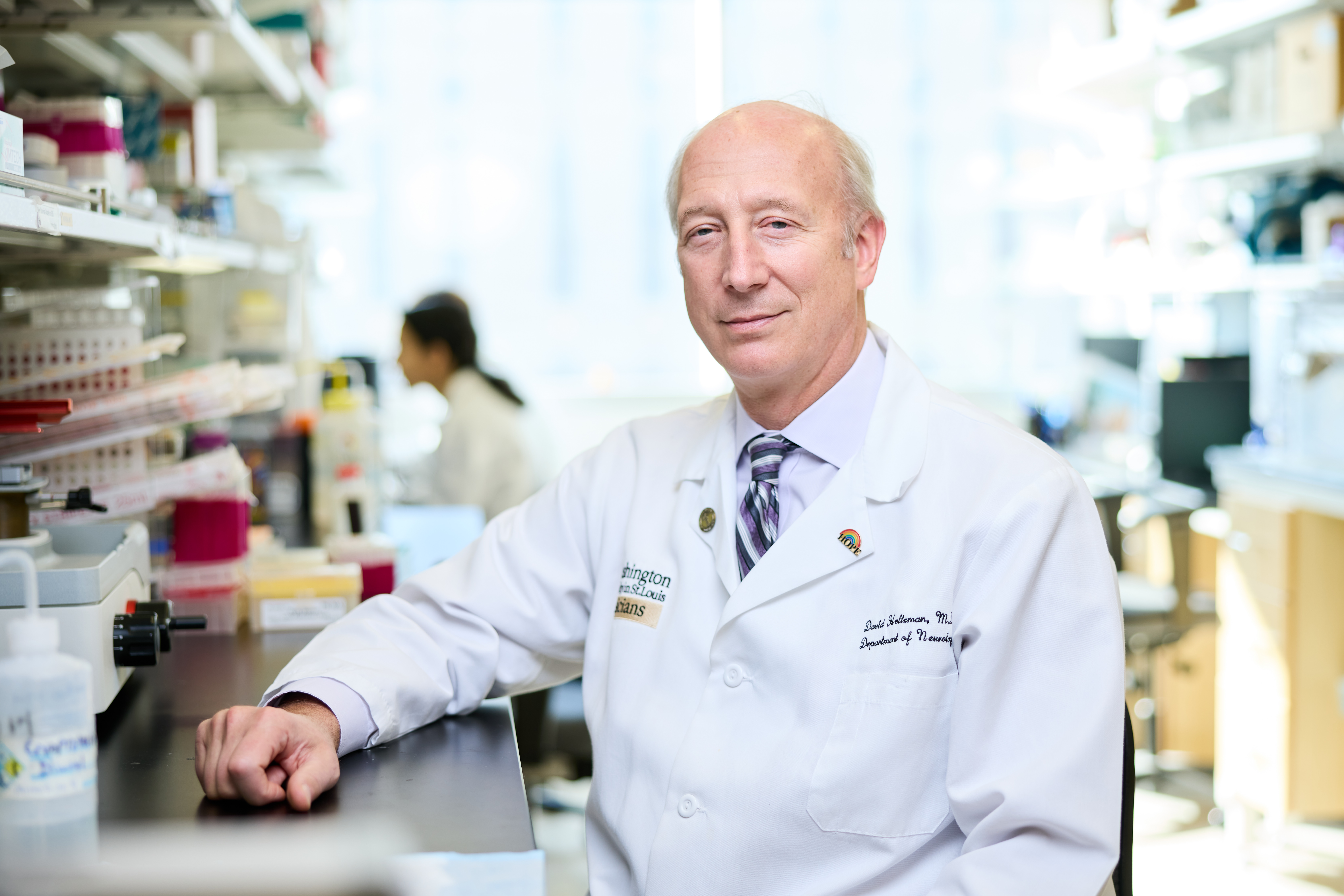 image of David Holtzman, MD, in white lab coat, sitting at a scientific laboratory bench